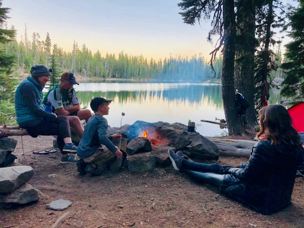 a family gathered around a campfire by a lake, with trees surrounding the area